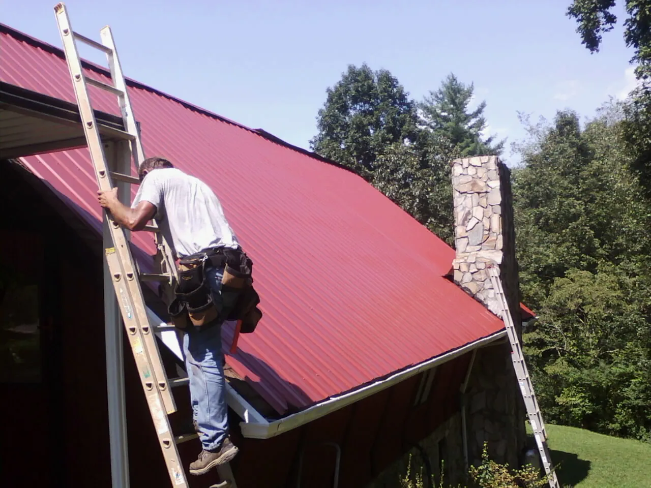 Roofer on a ladder beside a red metal roof and stone chimney — Soffit & Fascia Repair by Roofing Brothers in Stoneham